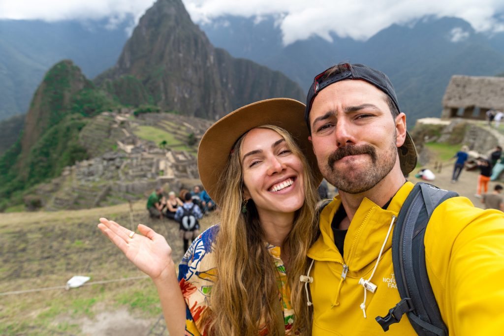 Ale e Duda sorrindo em uma selfie em Machu Picchu, com a montanha Huayna Picchu e as ruínas incas ao fundo.