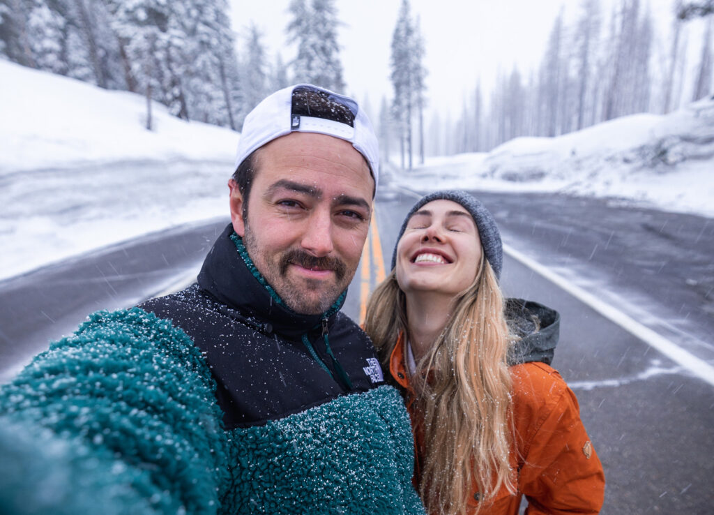 Ale e Duda sorrindo em selfie durante nevasca, em estrada cercada por pinheiros cobertos de neve