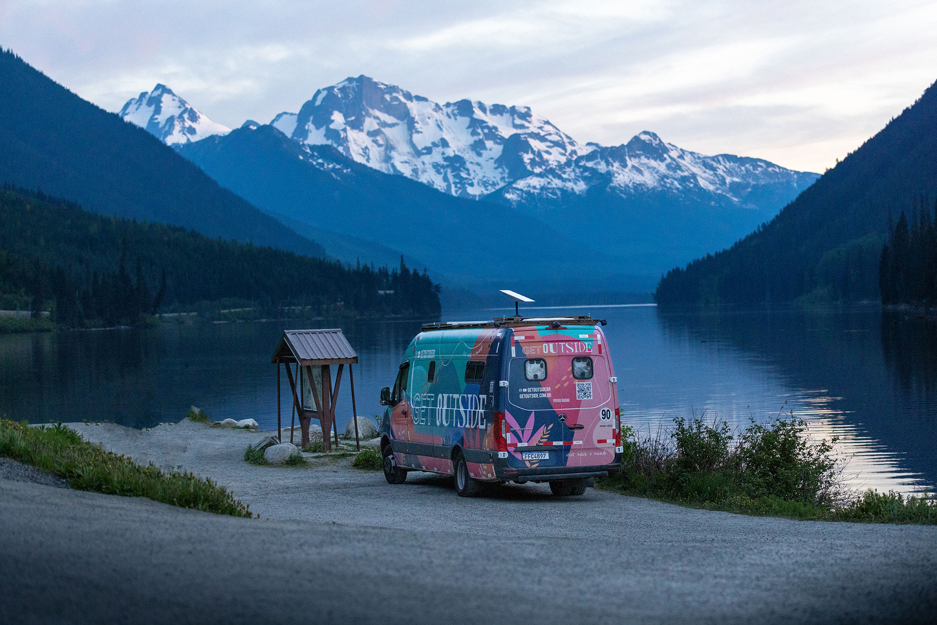 Van adesivada da Get Outside estacionada à beira de um lago, com montanhas nevadas ao entardecer