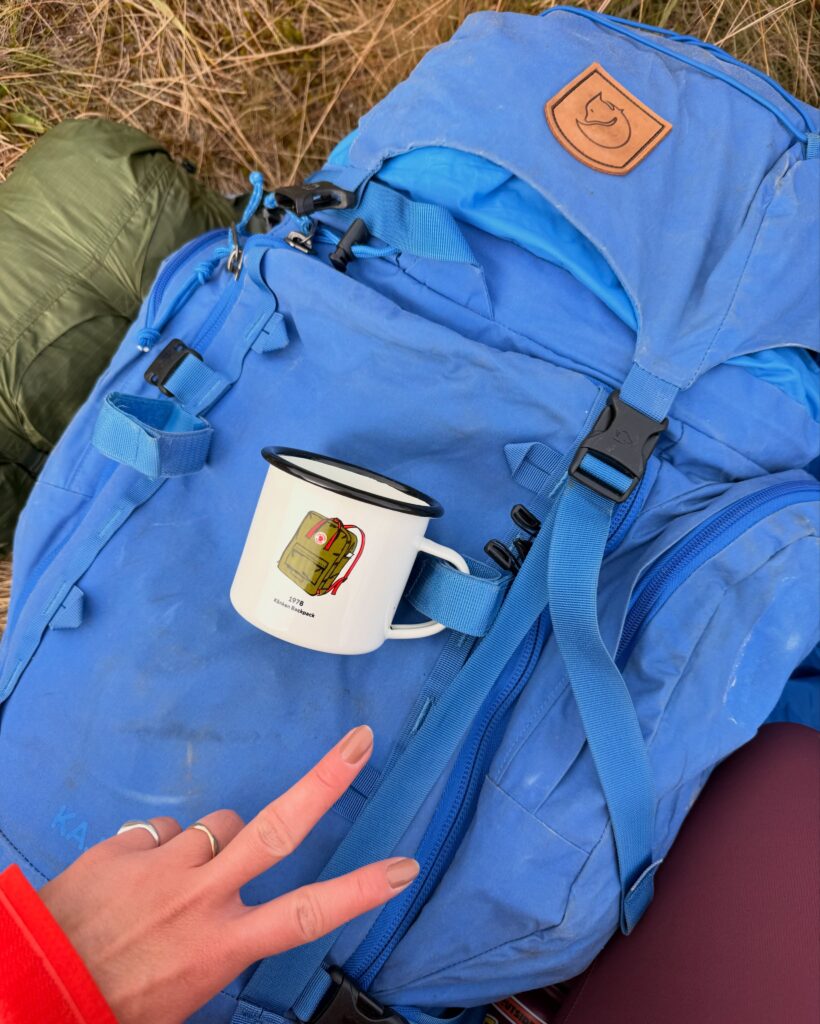 Mochila cargueira Fjällräven utilizada durante trekking, com caneca da marca presa ao equipamento para uso em viagens e aventuras ao ar livre.